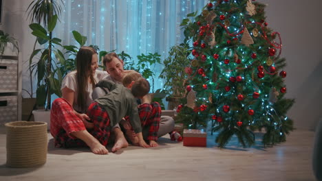 A-family-with-two-children-sit-together-at-the-Christmas-tree-and-cuddle.-Love-and-family-happiness-on-Christmas-Eve.-Father-and-two-sons-cuddle-and-look-at-Christmas-tree-in-their-living-room.-High-quality-4k-footage